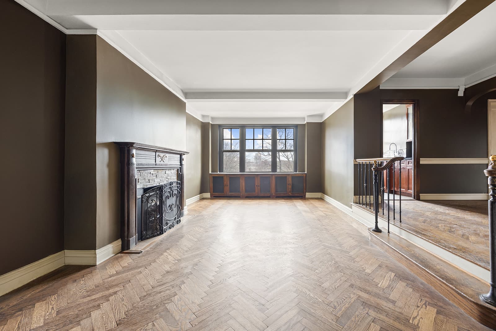 A long living room with herringbone floors, a carved fireplace and windows onto Riverside Park